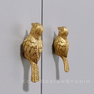 May include: Two gold bird-shaped cabinet door pulls. The birds are facing opposite directions and have detailed feather patterns.