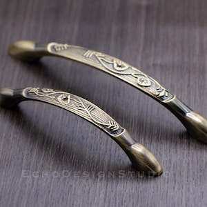 Könnte beinhalten: Zwei antike Messing-Schrankgriffe mit einem verzierten Blumenmuster. Die Griffe sind unterschiedlich groß und haben eine leicht gebogene Form.