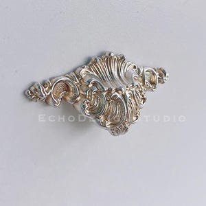 Può includere: Pomello per cassetto argentato e ornato con un design simmetrico e dettagliato. Il pomello è montato su una superficie bianca, con il testo "ECHOD STUDIO" visibile. Un elemento hardware decorativo.