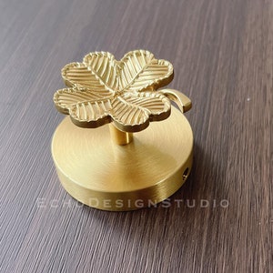 Puede incluir: Un gancho de metal dorado con un diseño de trébol de cuatro hojas. El gancho está montado en una base redonda.