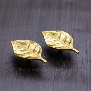 Könnte beinhalten: Zwei goldene, blattförmige Schrankknöpfe mit strukturierter Oberfläche.