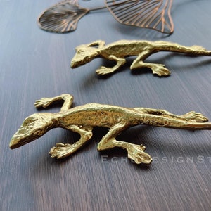 May include: Two gold-colored lizard figurines on a dark wood surface. The lizards are detailed with textured scales and have a realistic appearance.