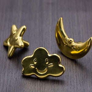 May include: Three gold-colored metal ornaments in the shape of a star, a crescent moon, and a cloud with a smiley face.