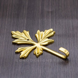 Puede incluir: Un colgador de pared dorado en forma de hoja. El colgador tiene un diseño curvo y está hecho de metal. La hoja tiene vetas detalladas y un pequeño agujero en el centro. El colgador está diseñado para colgar objetos.