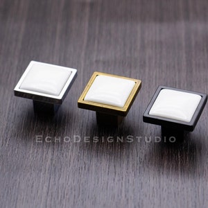 Puede incluir: Tres pomos cuadrados para armarios con inserciones de cerámica blanca. Los pomos se muestran en tres acabados: cromo, oro y negro.