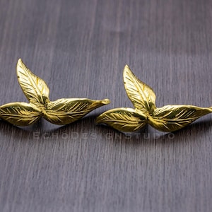 Puede incluir: Un par de pendientes de hoja de oro. Los pendientes están hechos de metal y tienen un diseño delicado y orgánico.