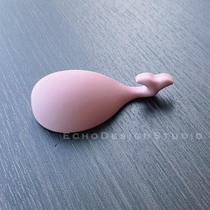 Puede incluir: Un reposacucharas de silicona rosa en forma de ballena. El reposacucharas está sobre una superficie gris oscura.
