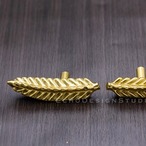 May include: Two gold-colored leaf-shaped cabinet pulls. Each pull has a central post for mounting and a detailed leaf design. The pulls are made of metal and are displayed on a dark wood surface.