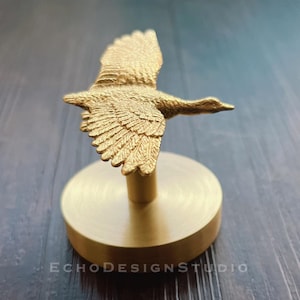 以下が含まれることがあります： 広げた翼を持つ金色の金属製の鳥の置物で、丸い金色の台座の上に置かれています。鳥は飛行中で、翼を広げています。台座は滑らかで磨かれています。