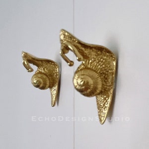 May include: Two gold snail-shaped wall hooks. The larger snail is on the right and the smaller snail is on the left. The snails are facing the right side of the image.