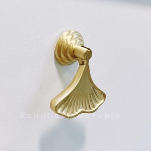 May include: Gold-toned ginkgo leaf-shaped cabinet pull with a decorative twisted knob. The pull is mounted on a white surface.