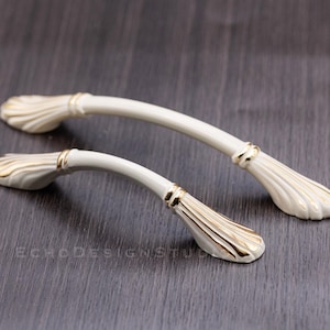 Op de afbeelding: Twee crèmekleurige kastgrepen met een schelpmotief en gouden accenten. De gebogen grepen hebben een sierlijke stijl. De hardware staat tegen een donkere houtnerf achtergrond.