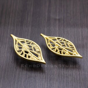 May include: A pair of gold leaf-shaped earrings with a delicate, intricate design. The earrings are made of metal and have a shiny finish.