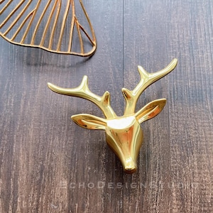 Könnte beinhalten: Eine goldfarbene Hirschkopfskulptur mit detailliertem Geweih. Die Skulptur besteht aus einem glänzenden, reflektierenden Material. Das Bild zeigt auch ein goldfarbenes, abstraktes Metallkunstwerk. Der Hintergrund ist eine dunkle Holzoberfläche.