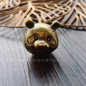 Puede incluir: Una detallada figura de cabeza de panda de color bronce. El panda tiene rasgos faciales visibles y orejas pequeñas y redondeadas. La figura está colocada sobre una superficie de madera oscura y texturizada, con un diseño de hojas doradas en el fondo.