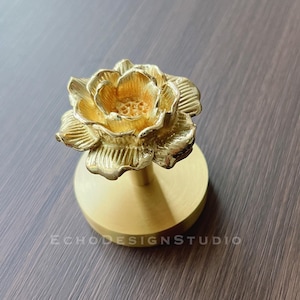 May include: A gold-coloured metal rose flower design on a round base. The rose is detailed with intricate petals and a centre.