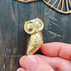 May include: A gold-colored owl figurine with detailed features. The owl is standing on a dark surface.
