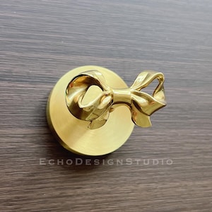 May include: A gold-toned metal door knob with a bow design. The knob is mounted on a round base.