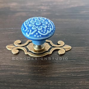 May include: A round, light blue ceramic knob featuring a white embossed floral pattern, mounted on an ornate, antiqued brass-toned metal backplate with a decorative scroll design. The text ECHODESIGNSTUDIO is visible on the metal base.