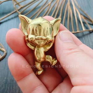 May include: A gold-coloured figurine of a cartoon cat in a playful pose, with its arms raised and one leg bent. The cat has large ears and a smiling expression. The figurine is held in a hand.