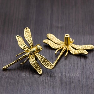 May include: Two gold-toned dragonfly drawer pulls with intricate details. The pull on the left has a long, slender body and delicate wings. The pull on the right has a shorter body and wider wings. Both pulls have a textured surface.