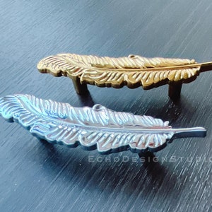 Könnte beinhalten: Zwei federförmige Schrankgriffe, einer golden und einer silbern, auf einer dunklen Holzoberfläche.