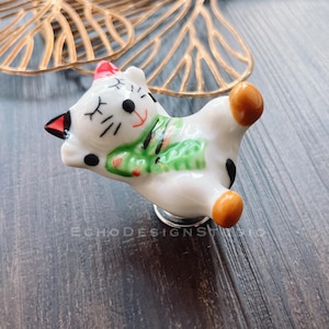 May include: White ceramic cat door knob with green and red details. The cat is lying down with its eyes closed. The knob is attached to a silver base.