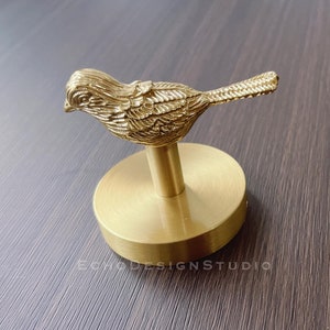 May include: A gold metal bird-shaped hook with a round base. The hook is designed to hang items on a wall.