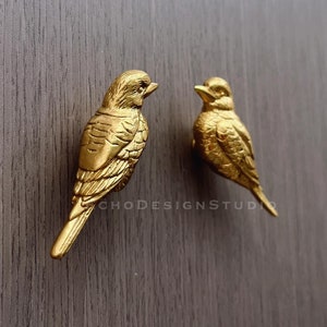 May include: Two gold bird-shaped cabinet knobs, facing each other, mounted on a wooden surface.