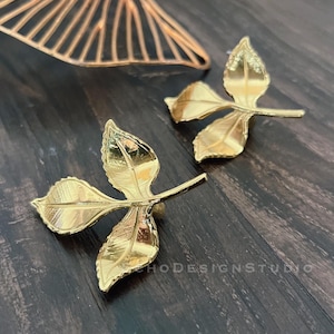 Könnte beinhalten: Zwei goldene Ohrringe in Blattform mit einem filigranen Design. Die Ohrringe sind aus Metall und haben eine glänzende Oberfläche.