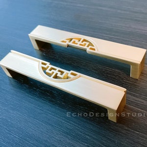 Peut inclure: Deux poignées de cabinet dorées avec un motif géométrique décoratif. Les poignées sont rectangulaires avec une découpe au centre.