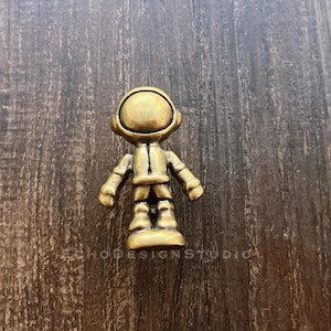 May include: A small, gold-colored astronaut figurine. The astronaut is wearing a helmet and a spacesuit. The figurine is standing on a small base. The background is a dark wood surface. The text "ECHODESIGNSTUDIO" is visible.
