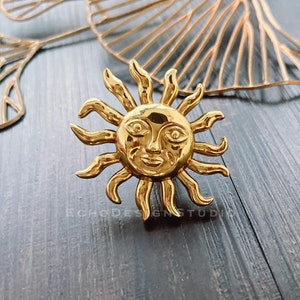 Puede incluir: Un anillo de sol dorado con una cara y rayos. El anillo está sobre una superficie de madera oscura.