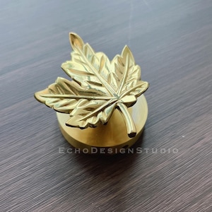 May include: A gold-colored maple leaf bottle stopper. The leaf is detailed and sits on a round, gold-colored base.