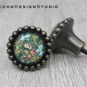 Könnte beinhalten: Zwei antike Messing-Schrankknöpfe mit floralem Design. Der Knopf links hat einen runden, abgeschrägten Glaseinsatz mit einem floralen Muster in den Farben Blau, Grün, Orange und Rot. Der Knopf rechts ist ein traditioneller Stil mit einem geriffelten Sockel.
