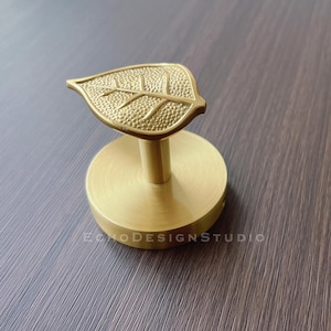 May include: A gold leaf-shaped hook with a round base. The hook is made of metal and has a textured surface.