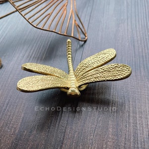 Può includere: Un decoro da parete a forma di libellula in metallo dorato con dettagli intricati. La libellula è posata su una superficie in legno scuro.