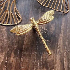 Könnte beinhalten: Eine goldfarbene Libellen-Wanddekoration mit filigranen Details. Die Libelle ist auf einer dunklen Holzoberfläche montiert.