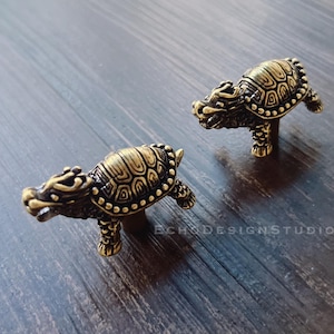 Könnte beinhalten: Zwei antike schildkrötenförmige Schrankknöpfe aus Messing. Die Knöpfe weisen detaillierte Schalenmuster und drachenartige Köpfe auf. Die Knöpfe befinden sich auf einer dunklen Holzoberfläche. Der Text "ECHO DESIGN STUDIO" ist sichtbar.