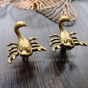 以下が含まれることがあります： アンティーク真鍮製のサソリ型キャビネットノブ2個セット。各ノブは、広げられた爪と湾曲した尾を持つ詳細なサソリのデザインが特徴です。暗い木製の表面に取り付けられており、家具にユニークなタッチを加えるのに最適です。