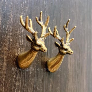 May include: Two brass-colored deer head wall decor pieces. Each deer head has detailed antlers and a textured surface. The deer heads are mounted on a dark wooden surface, creating a rustic aesthetic.