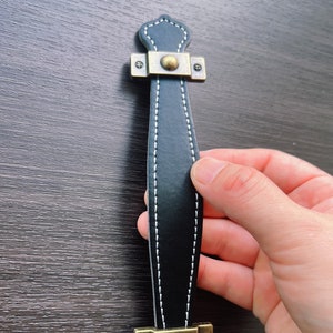 May include: Black leather handle with white stitching and brass accents. The handle has a decorative top and bottom.