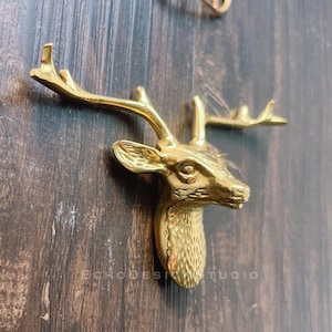 Op de afbeelding: Een gouden metalen hertenkop wandhaak. De hertenkop is gedetailleerd met een textuur en heeft een realistisch ontwerp.