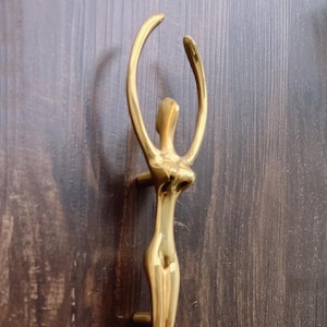 Puede incluir: Un tirador de puerta dorado con forma de figura humana estilizada con los brazos levantados. El tirador está montado en una puerta de madera marrón oscuro. El texto "ECHODESIGNSTUDIO" es visible en la parte inferior.