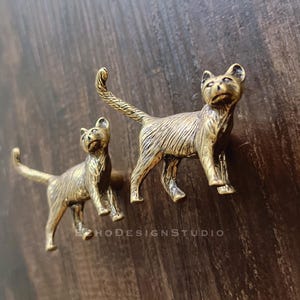May include: Two bronze cat figurines. The cats are in a standing position with their tails up. The figurines have detailed features and are set against a dark wood background. The text "ECHO DESIGN STUDIO" is visible.