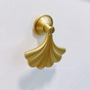 Może przedstawiać: Złoty uchwyt do szafki o kształcie wachlarza. Uchwyt jest przymocowany do białych drzwi szafki.