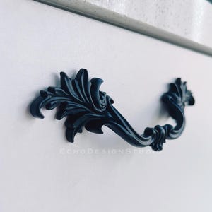 Może przedstawiać: Czarny, ozdobny uchwyt do szuflady z zakrzywionym uchwytem i detalami w kształcie liści. Uchwyt jest zamontowany na białych drzwiach szafki. Na drzwiach widoczny jest napis "ECHO DESIGN STUDIO".