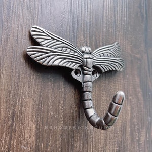 Puede incluir: Gancho de libélula plateado con un diseño detallado. El gancho tiene forma de libélula con alas y una cola rizada.