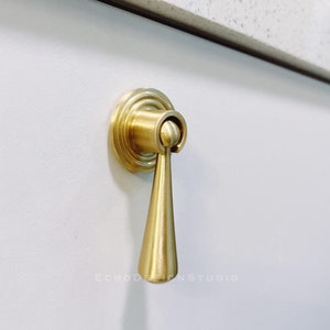 Puede incluir: Un pomo de armario dorado con una manija en forma de lágrima y una base redonda. El pomo está montado en una puerta de armario blanca.