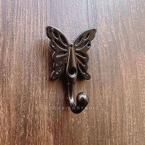 Peut inclure: Crochet en métal noir en forme de papillon avec un design décoratif. Le crochet est monté sur une surface en bois.